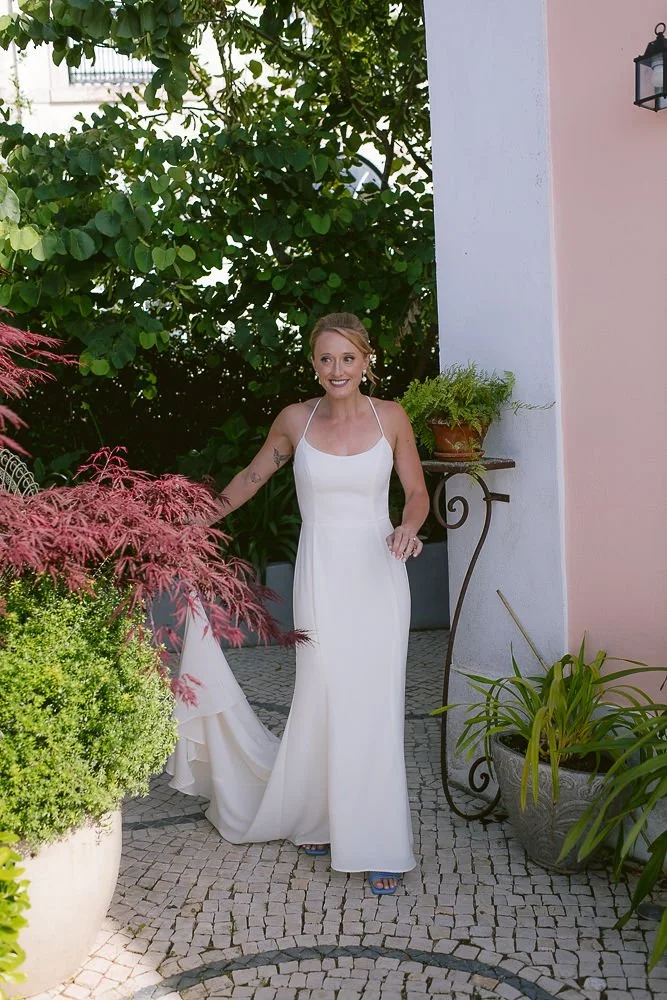 A woman in a white dress standing outdoors on a cobblestone pathway surrounded by lush green plants and pink flowers, with a pink wall and a black lantern in the background.