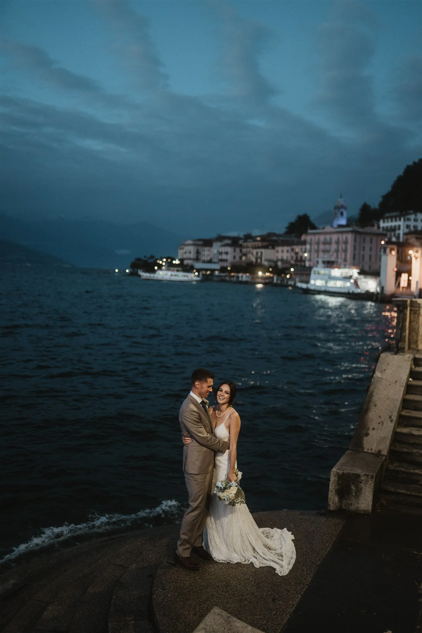 A bride and groom standing together at a waterfront during twilight, with a view of boats, buildings, and a cloudy sky in the background.