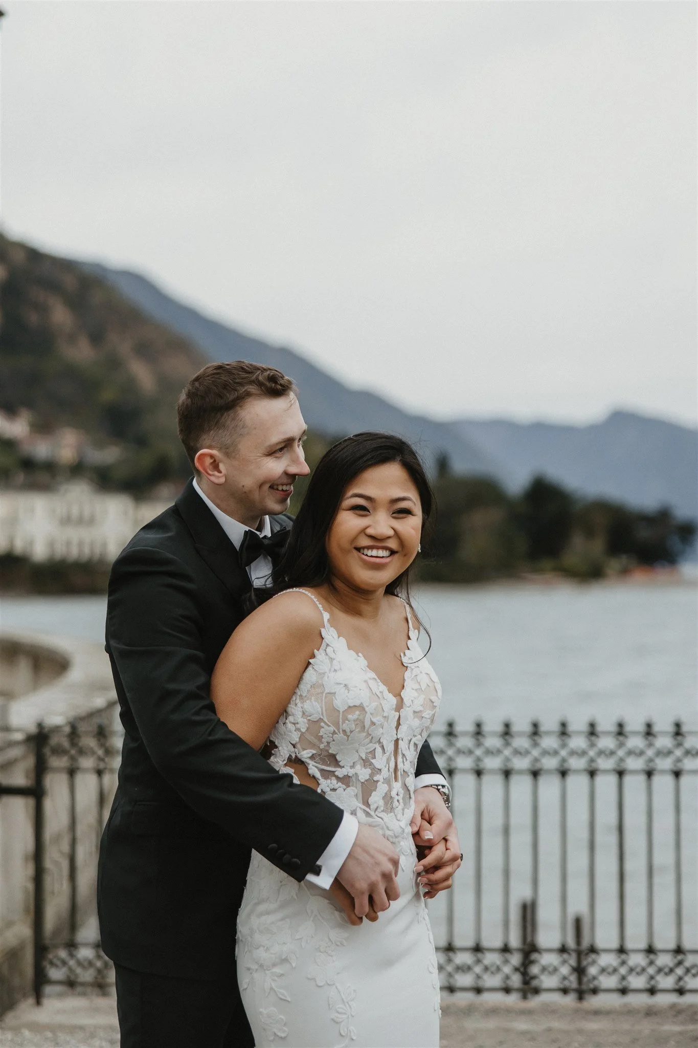 A smiling bride and groom in wedding attire standing by a waterfront with mountains in the background, during cloudy weather.