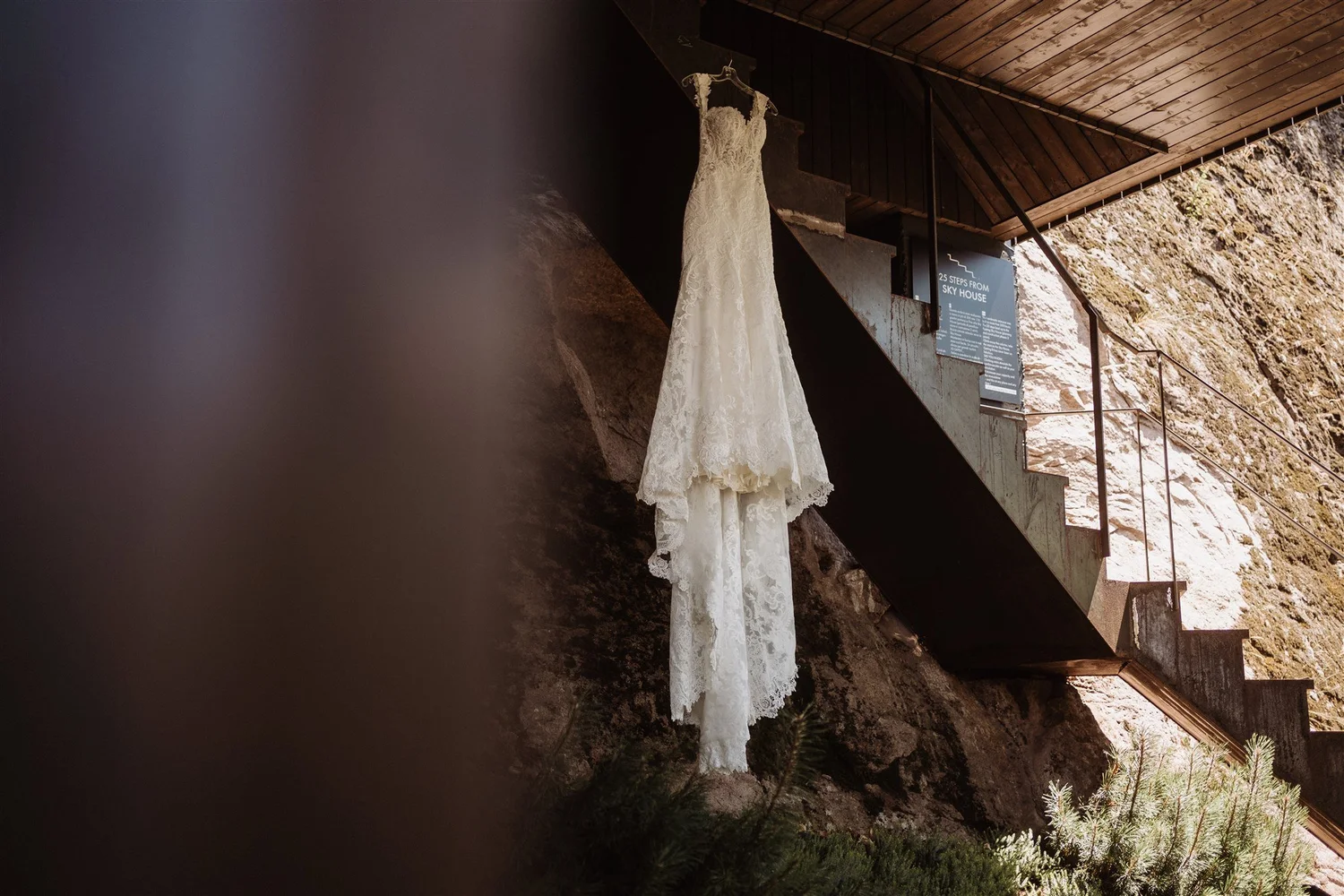 A white lace wedding dress hanging on a hanger from a black staircase railing against a rocky wall with a wooden ceiling in the background.