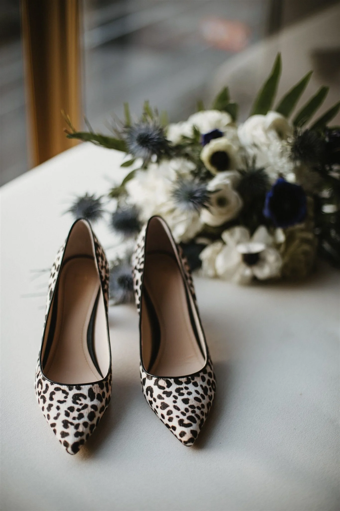 Pair of studded leopard print high heels on a white surface, with a blurry floral bouquet in the background.