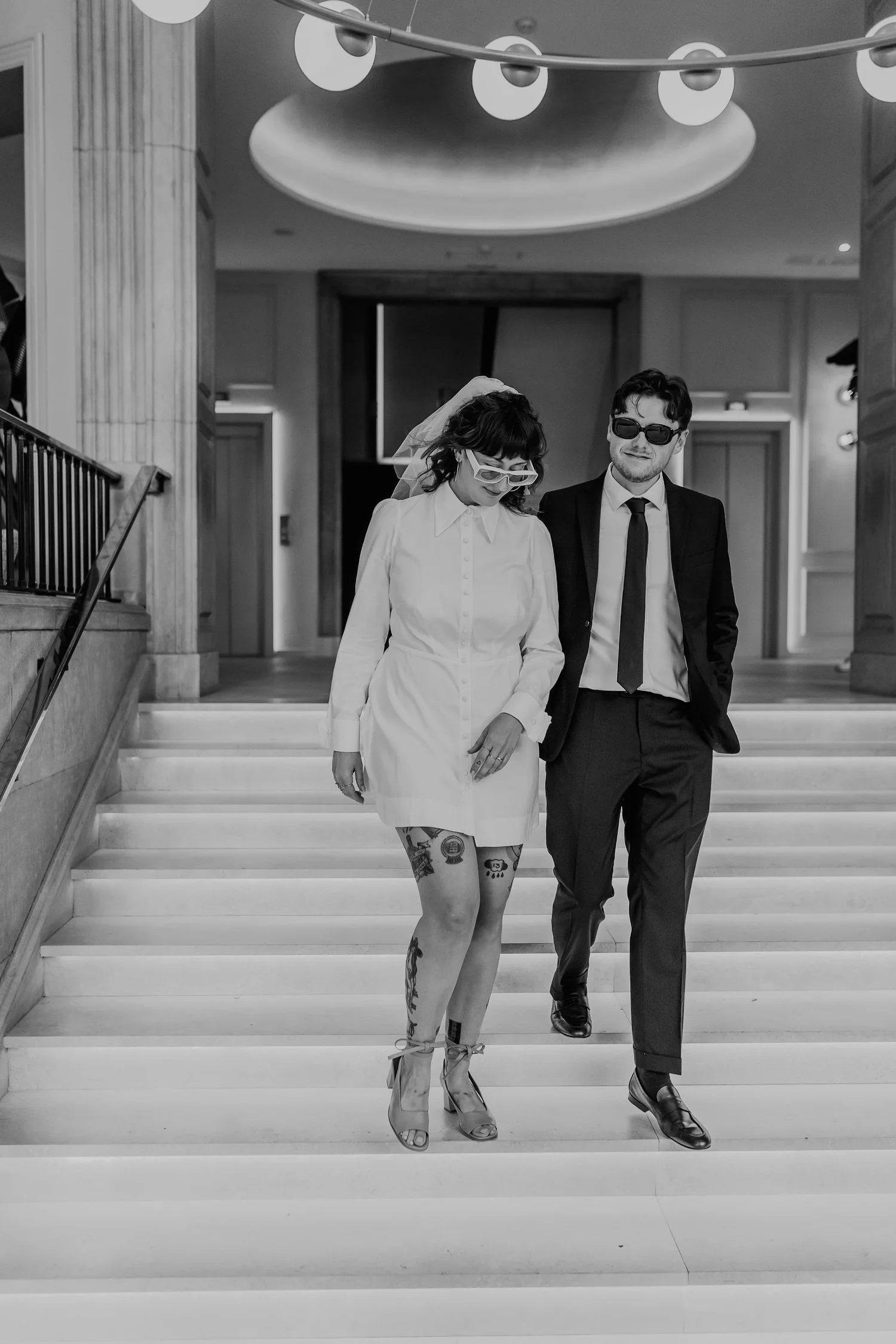 A black-and-white photo of a man and woman walking down a staircase in a modern indoor space. The woman is wearing a white dress, high heels, and glasses, with tattoos visible on her legs. The man is wearing a suit, tie, and sunglasses, with his hand