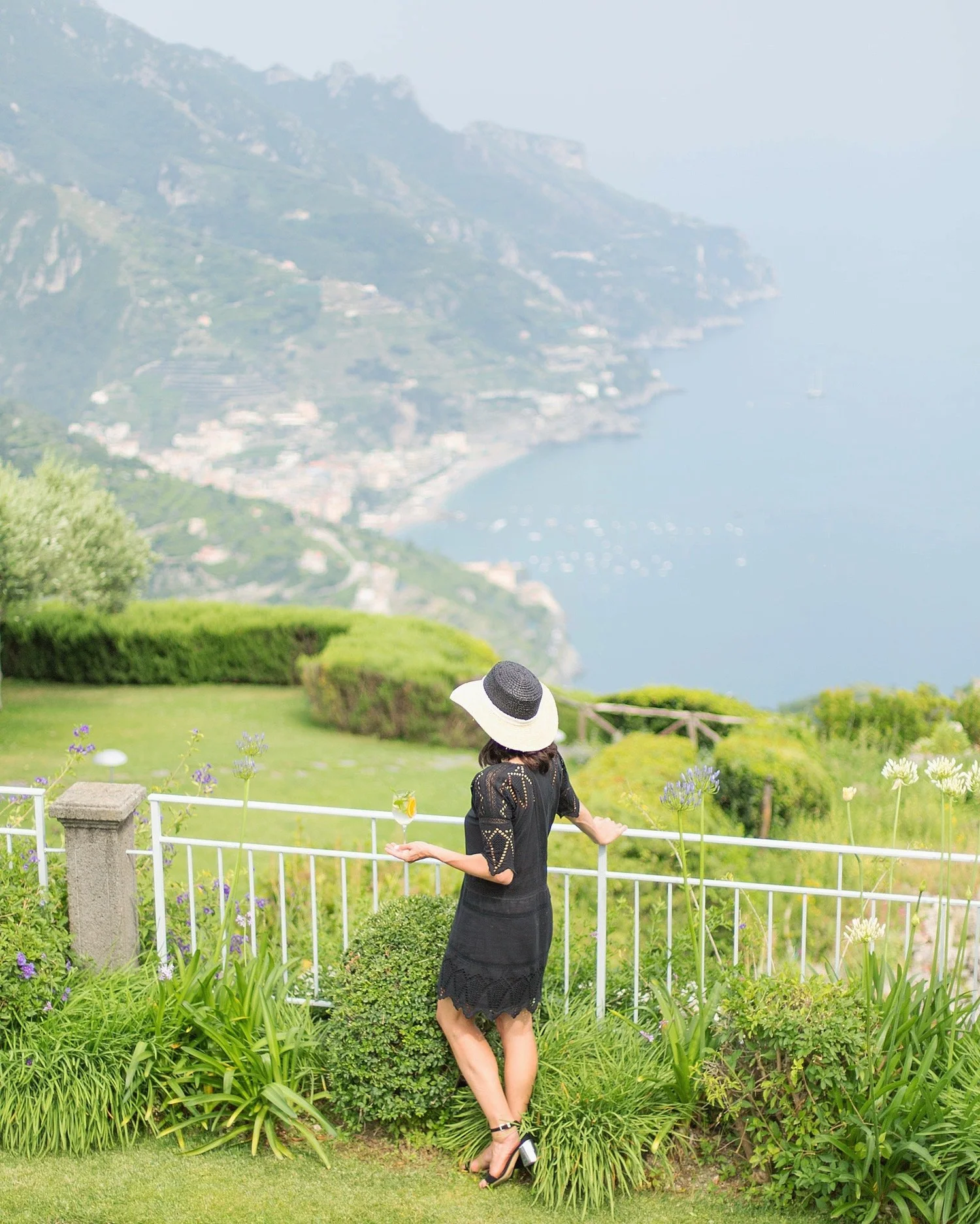 A woman wearing a black dress, black heels, and a wide-brimmed white hat with a black band stands in a garden with lush green plants and purple flowers, overlooking a scenic hillside and body of water in the background.
