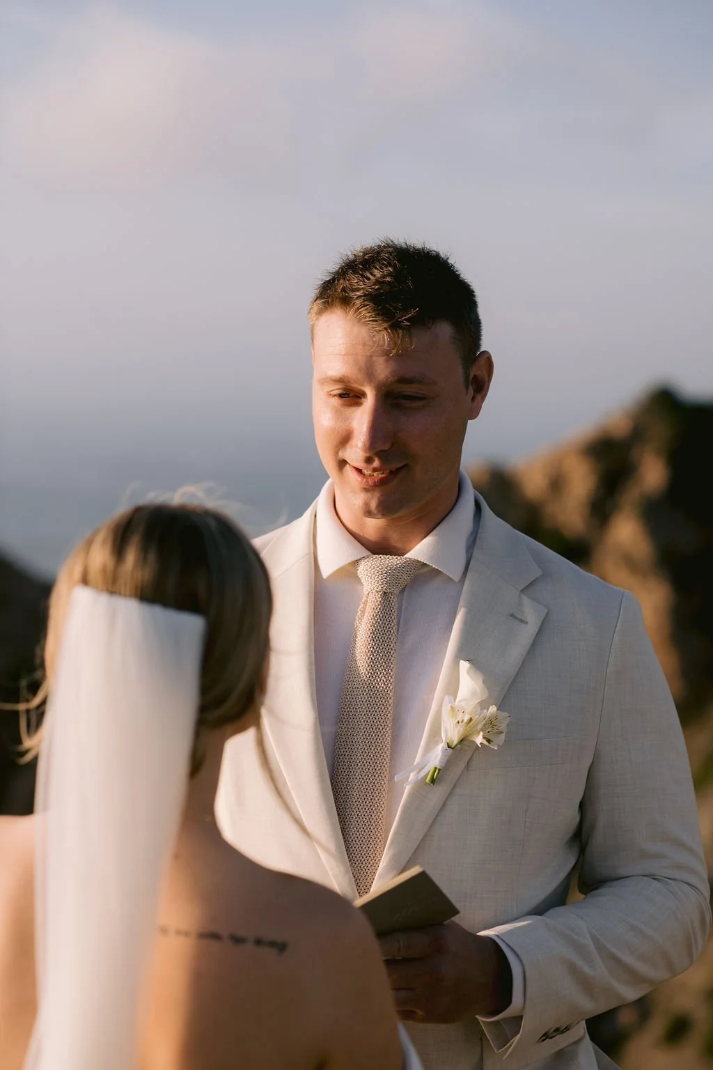A groom in a light-colored suit and tie looks at a bride with long hair and a veil during an outdoor wedding ceremony with rocky cliffs in the background.