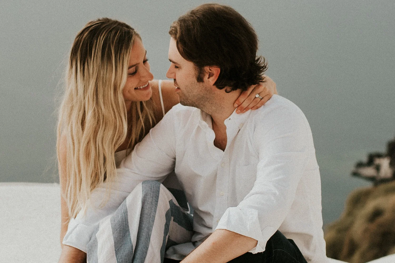 A woman with long blonde hair and light skin leaning close to a man with dark hair and light skin, both smiling and appearing affectionate, in a cozy indoor setting.