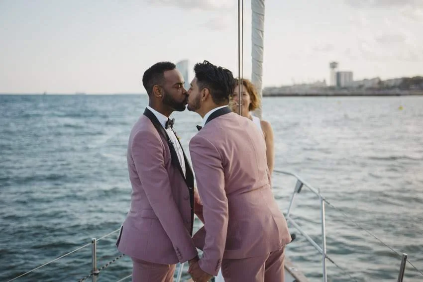 Two men in pink suits holding hands and touching noses on a boat, with a woman in the background near the water.