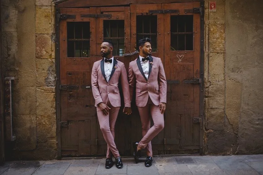 Two men in pink tuxedos with black lapels, black bow ties, and black dress shoes standing in front of a rustic wooden garage door, holding hands and looking in opposite directions.