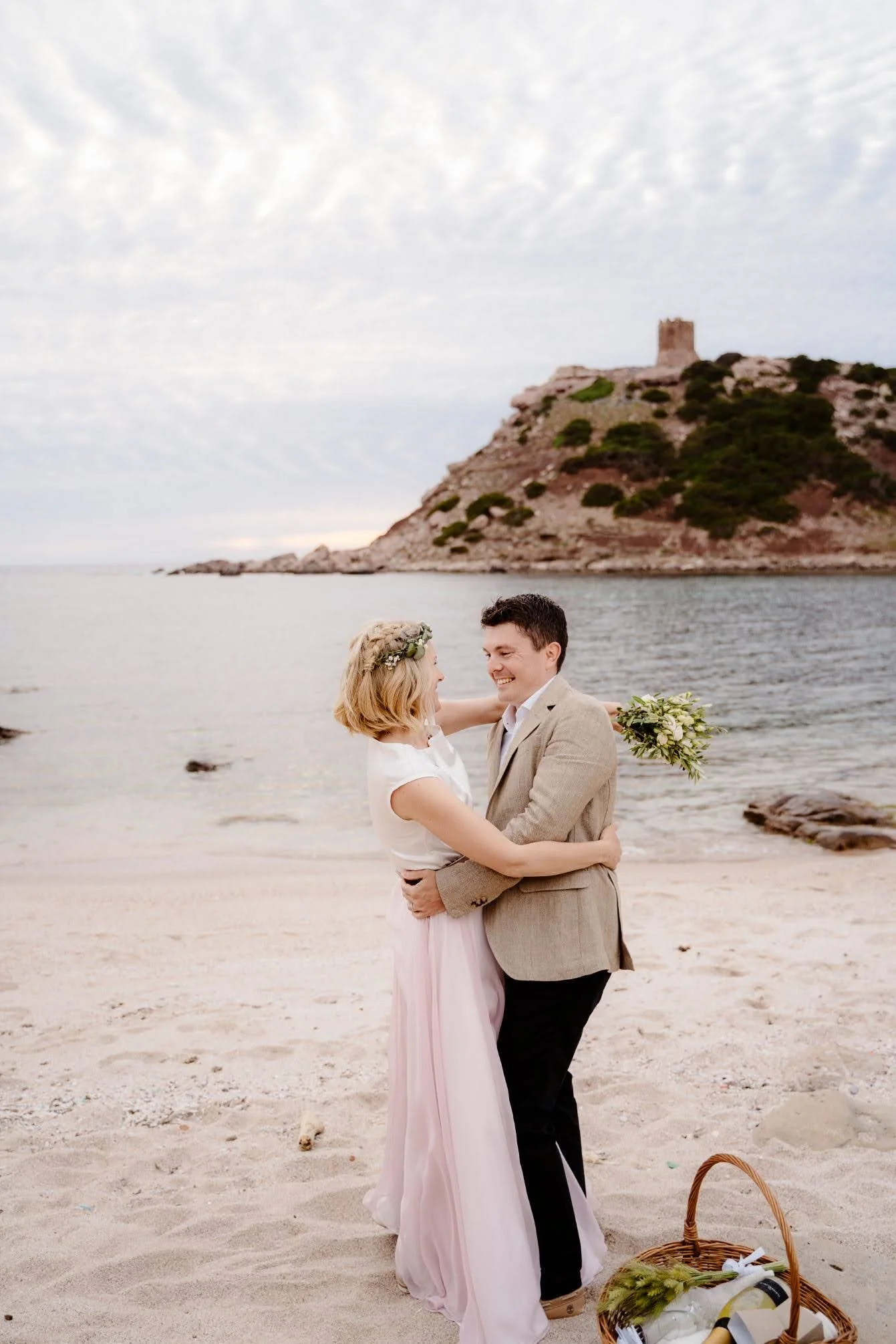 A couple dancing on the beach, with a castle on a hill in the background, during a cloudy day.