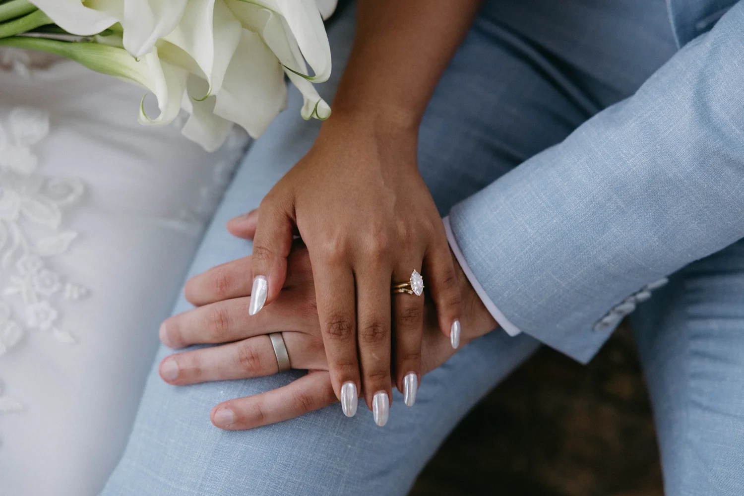 Close-up of a couple holding hands, focusing on their interlocked fingers and wedding rings, with a bouquet of white flowers nearby.