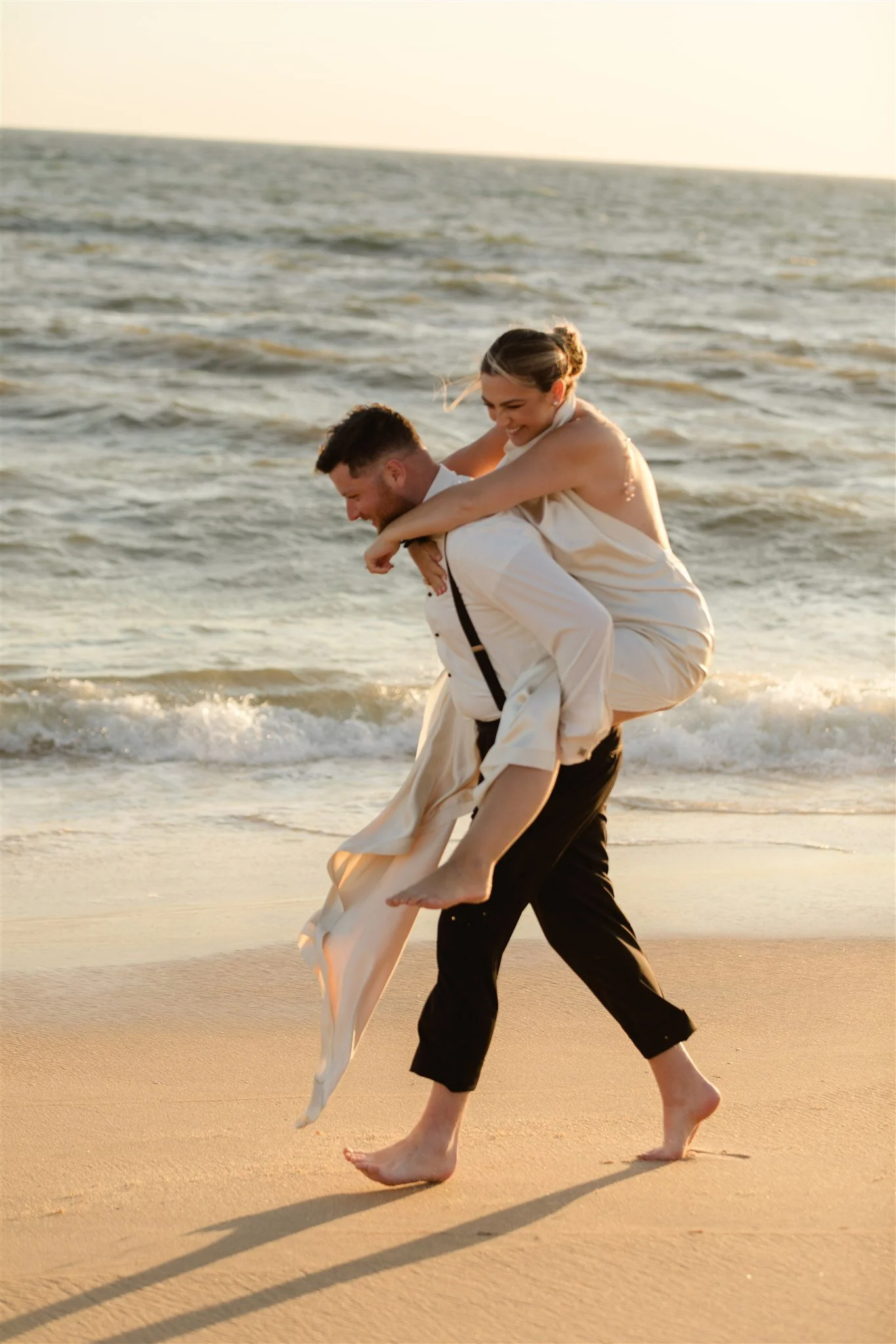 A couple on the beach during sunset, with the woman piggybacking on the man, both smiling and enjoying the moment, with the ocean and sky in the background.