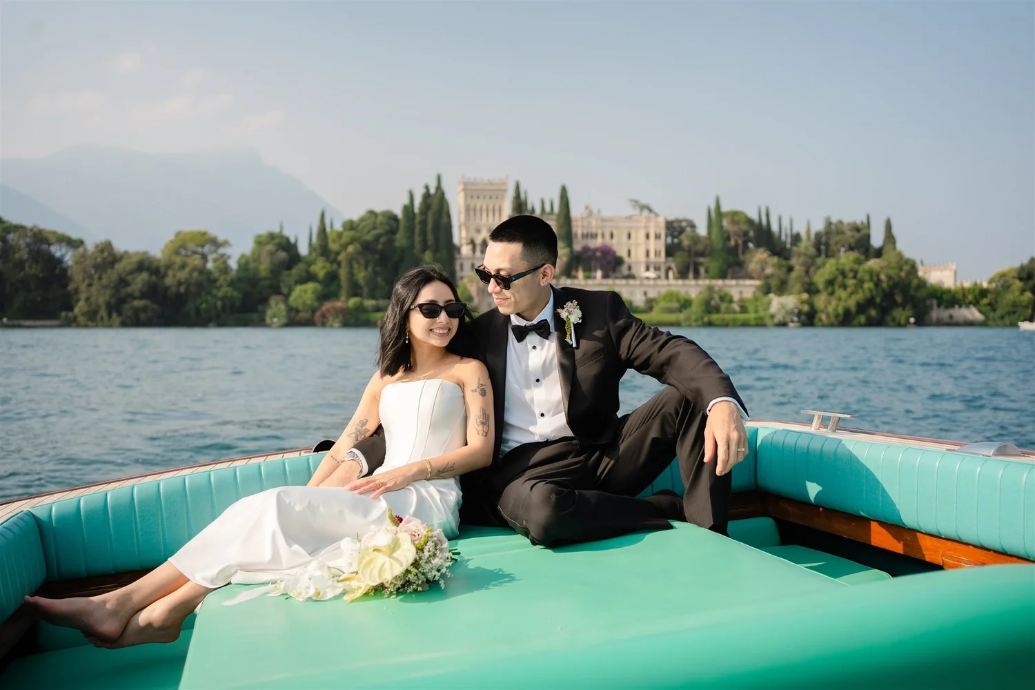 A happy couple dressed in wedding attire sitting on a boat on a lake, with a historical castle and lush green trees in the background.