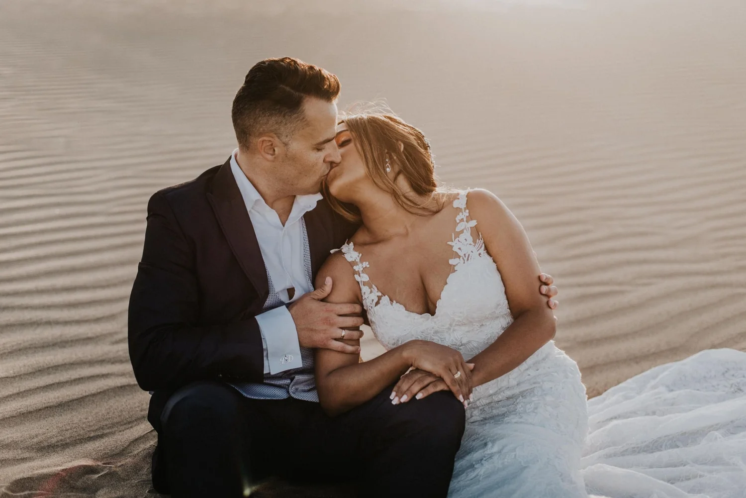 A couple, dressed in wedding attire, sharing a kiss on a sandy beach with calm waters in the background.
