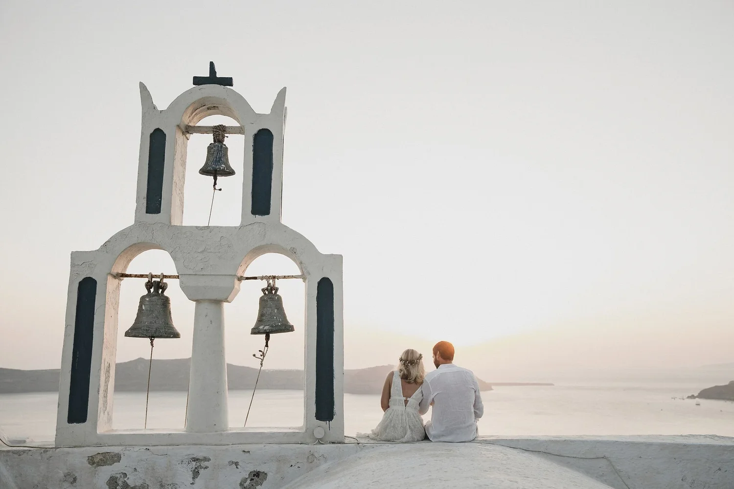 A white church bell tower with three bells, set outdoors on a white wall, overlooking a beach and ocean at sunset. A couple in wedding attire sit on the ground, facing away from the camera, near the tower.