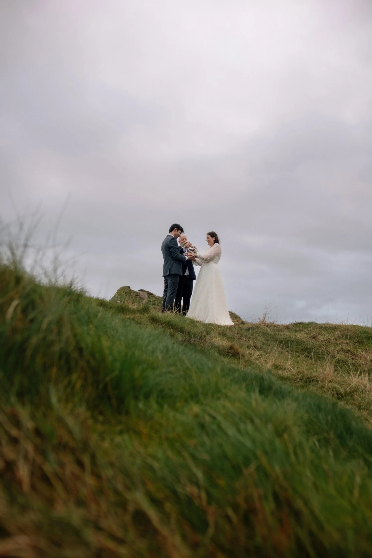 A couple dressed in wedding attire standing on a grassy hill under a cloudy sky, holding a small dog.