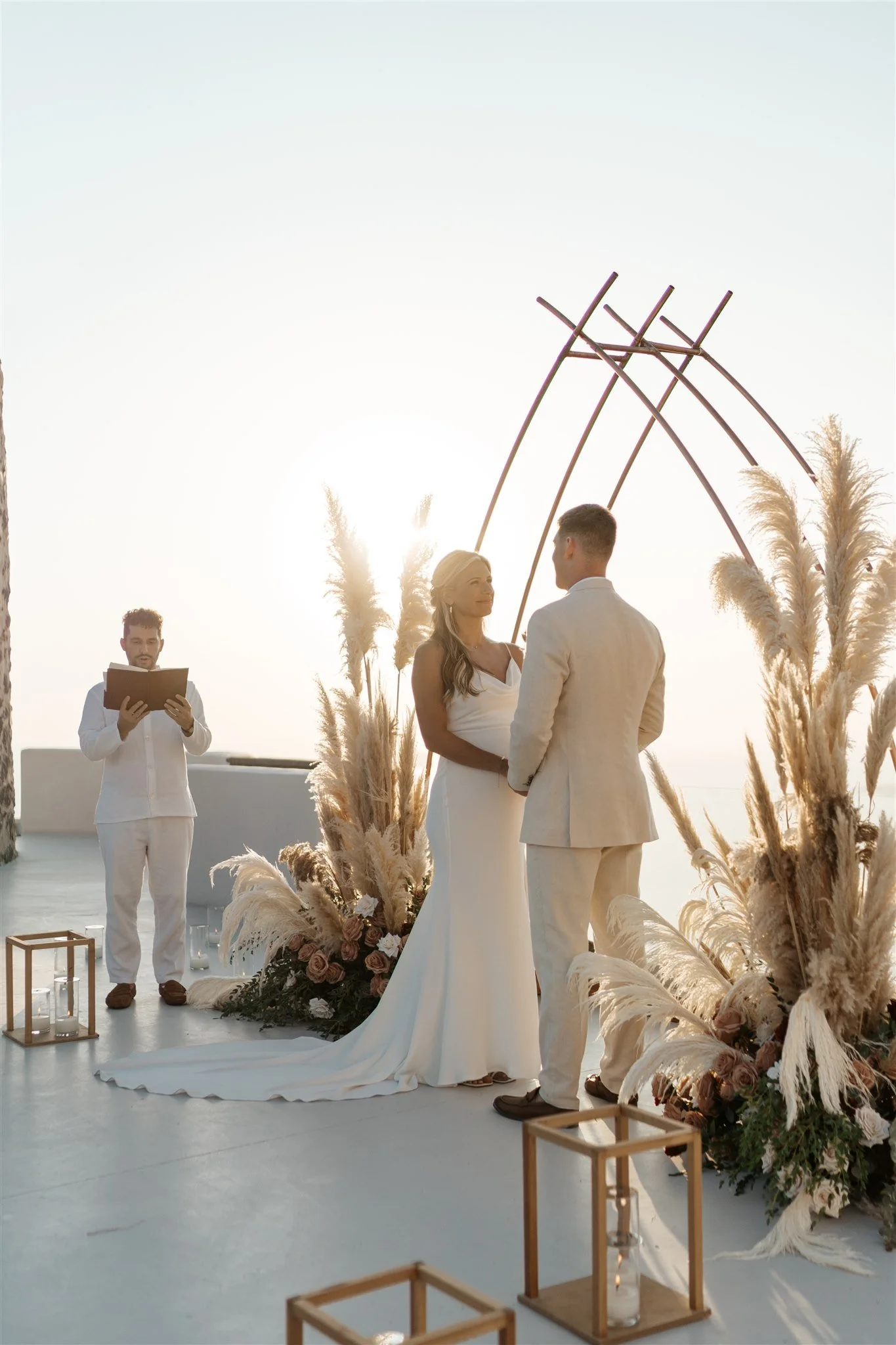 A couple getting married outdoors during sunset, with the bride wearing a white wedding gown and the groom in a light-colored suit, standing under a unique geometric wooden arch decorated with dried pampas grass. An officiant reads from a book nearby, and candles are placed in wooden lanterns on the ground, with floral arrangements at the base of the arch.