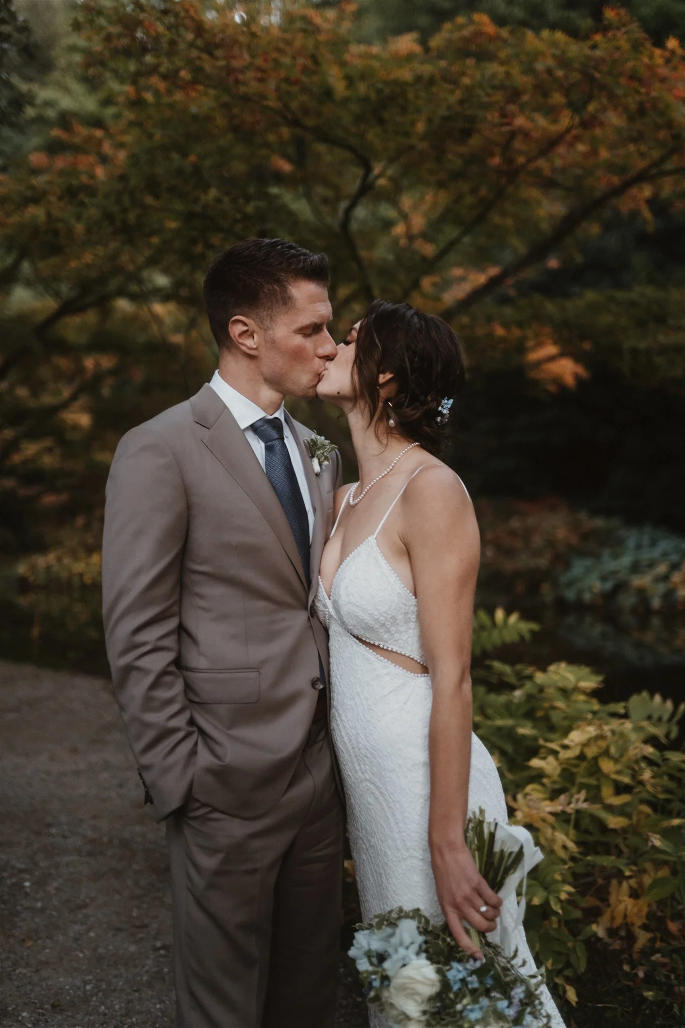 A newlywed couple sharing a kiss outdoors in front of autumn foliage. The groom is wearing a tan suit, and the bride is dressed in a white lace wedding gown holding a bouquet of flowers.