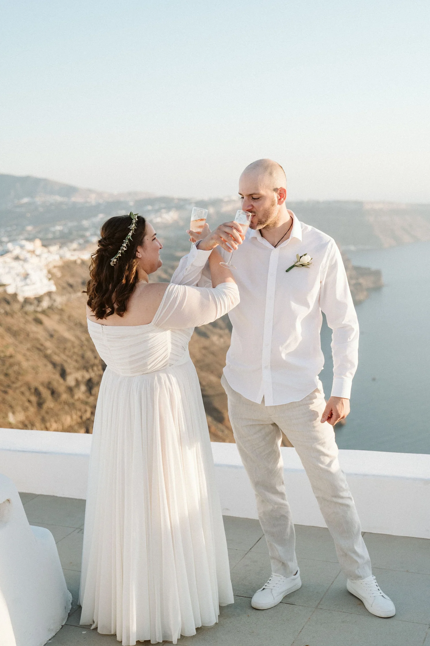 A couple dressed in white, celebrating with drinks, outdoors overlooking a coastline with hills and water in the background.