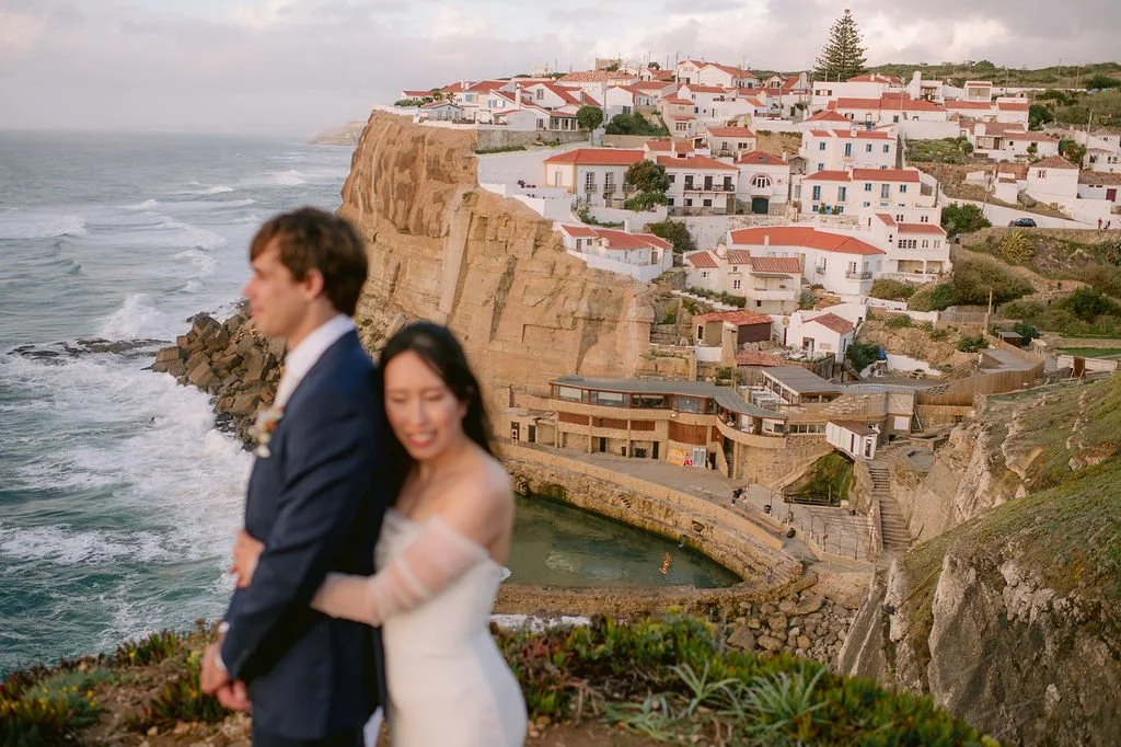 A couple dressed in wedding attire stands in the foreground with a coastal village and cliffs in the background.