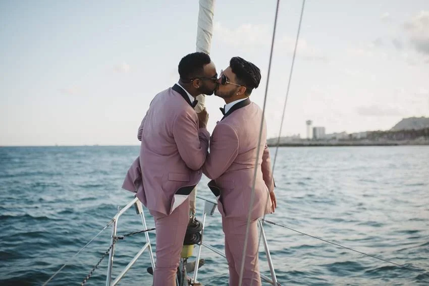Two men in pink tuxedos sharing a kiss on a boat with water and a distant shoreline in the background.
