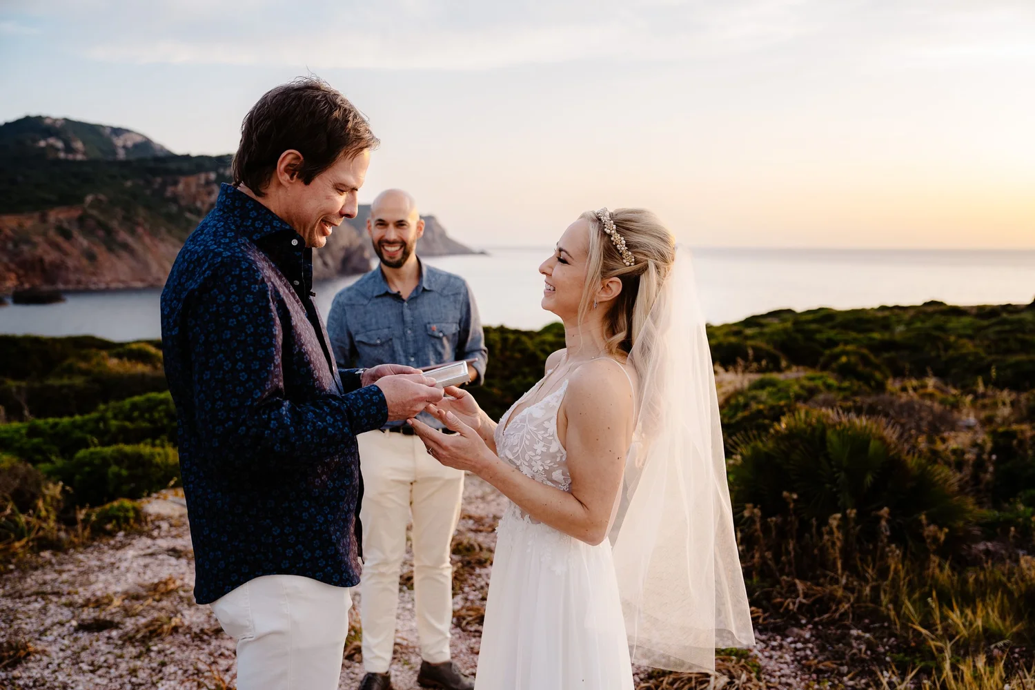 A wedding ceremony outdoors at sunset with a bride and groom exchanging rings, accompanied by a man standing behind them, in front of a scenic coastal landscape with cliffs and water.