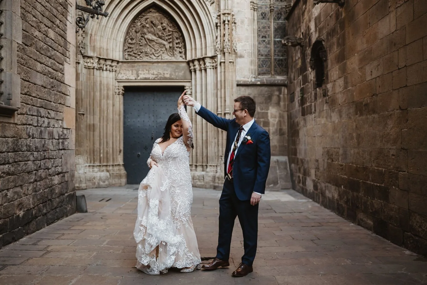 A bride and groom dancing together outside in a narrow, stone alleyway with an ornate archway and carved stone relief in the background. The bride wears a white lace wedding dress, and the groom is in a navy suit with a striped tie and boutonniere. T