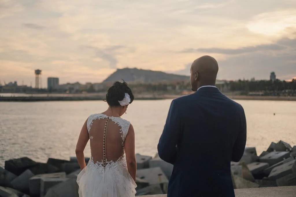 A bride and groom stand by a body of water during sunset, with city buildings and a hill in the background.