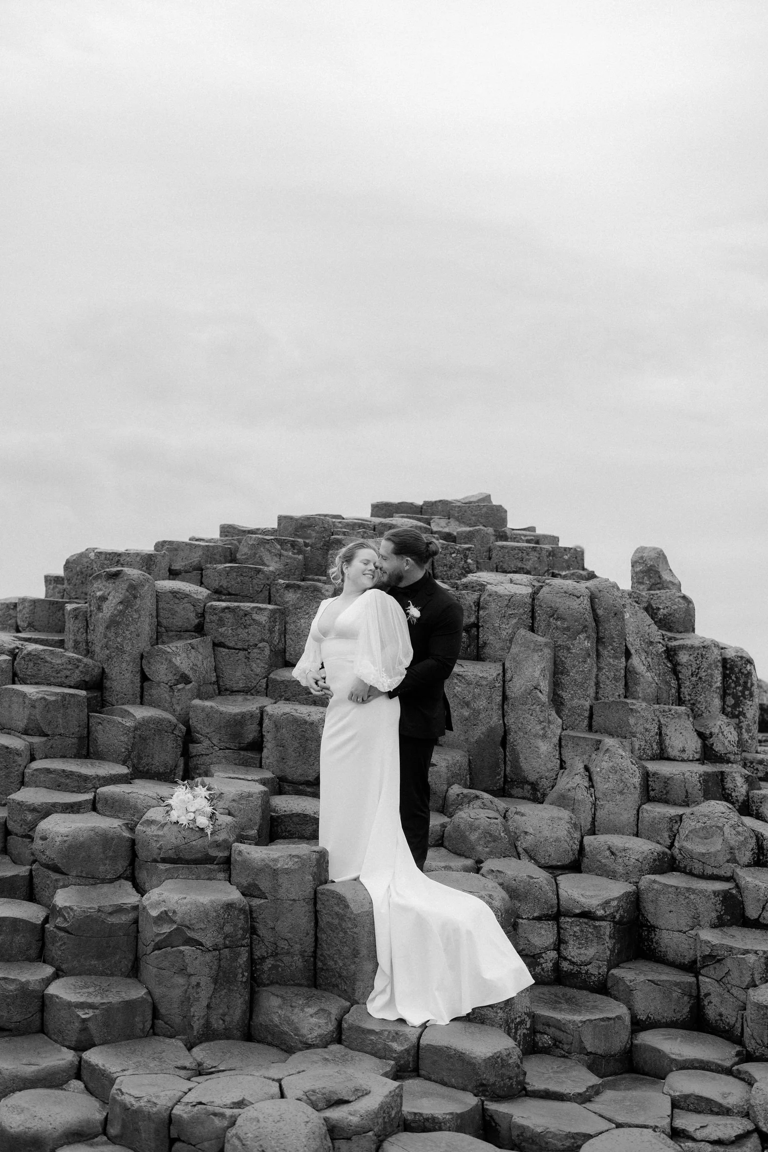 A bride and groom are standing on an arrangement of large rocks, with a cloudy sky above. The bride is in a white wedding gown, holding a bouquet of flowers in front of her, while the groom is in a dark suit, embracing her and smiling. They are sharing a tender moment.
