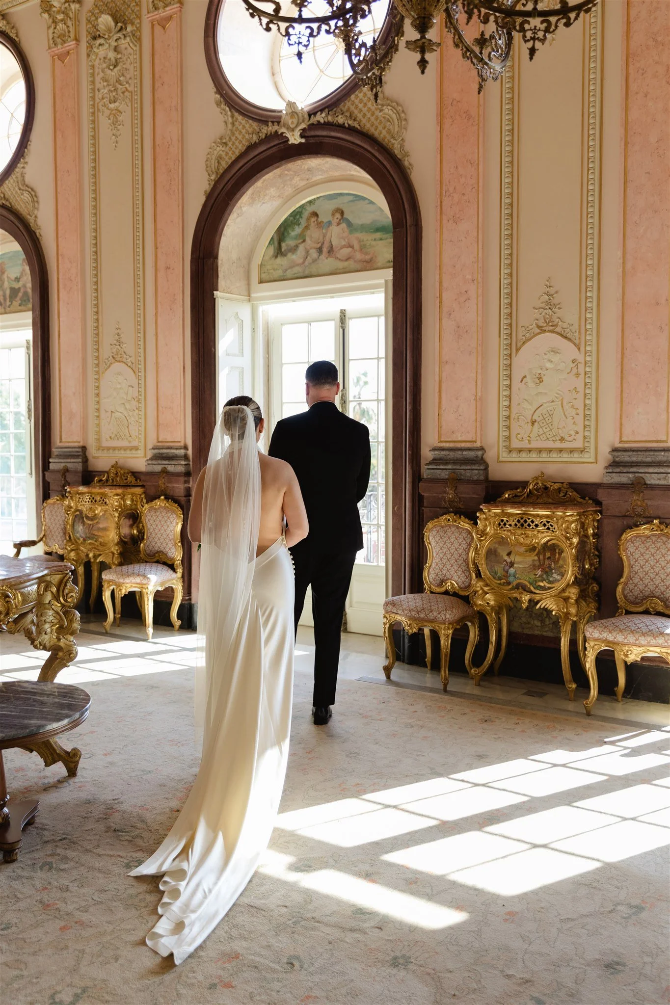 A bride in a white wedding dress with a veil, and a groom in a black suit, stand together in a lavishly decorated room with gold accents, ornate furniture, and large windows. They are facing away from the camera, looking toward the window.
