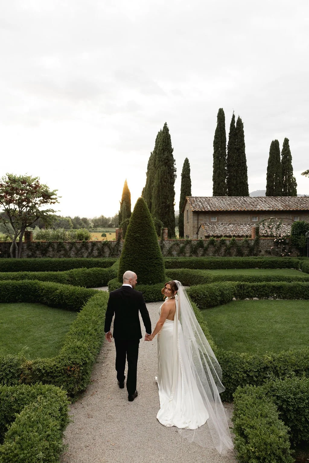 A bride and groom holding hands walk down a gravel garden path in front of neatly trimmed bushes and tall cypress trees in a scenic outdoor setting during sunset.