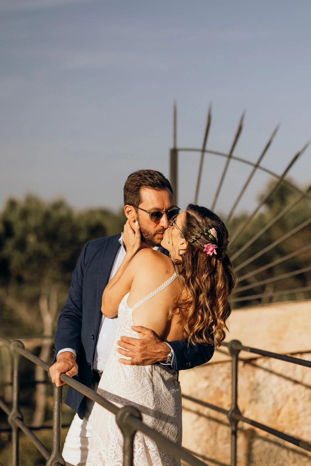 A couple sharing a romantic kiss outdoors, with the woman wearing a white lace dress and the man in a navy suit and sunglasses, on a bridge with a scenic natural background.