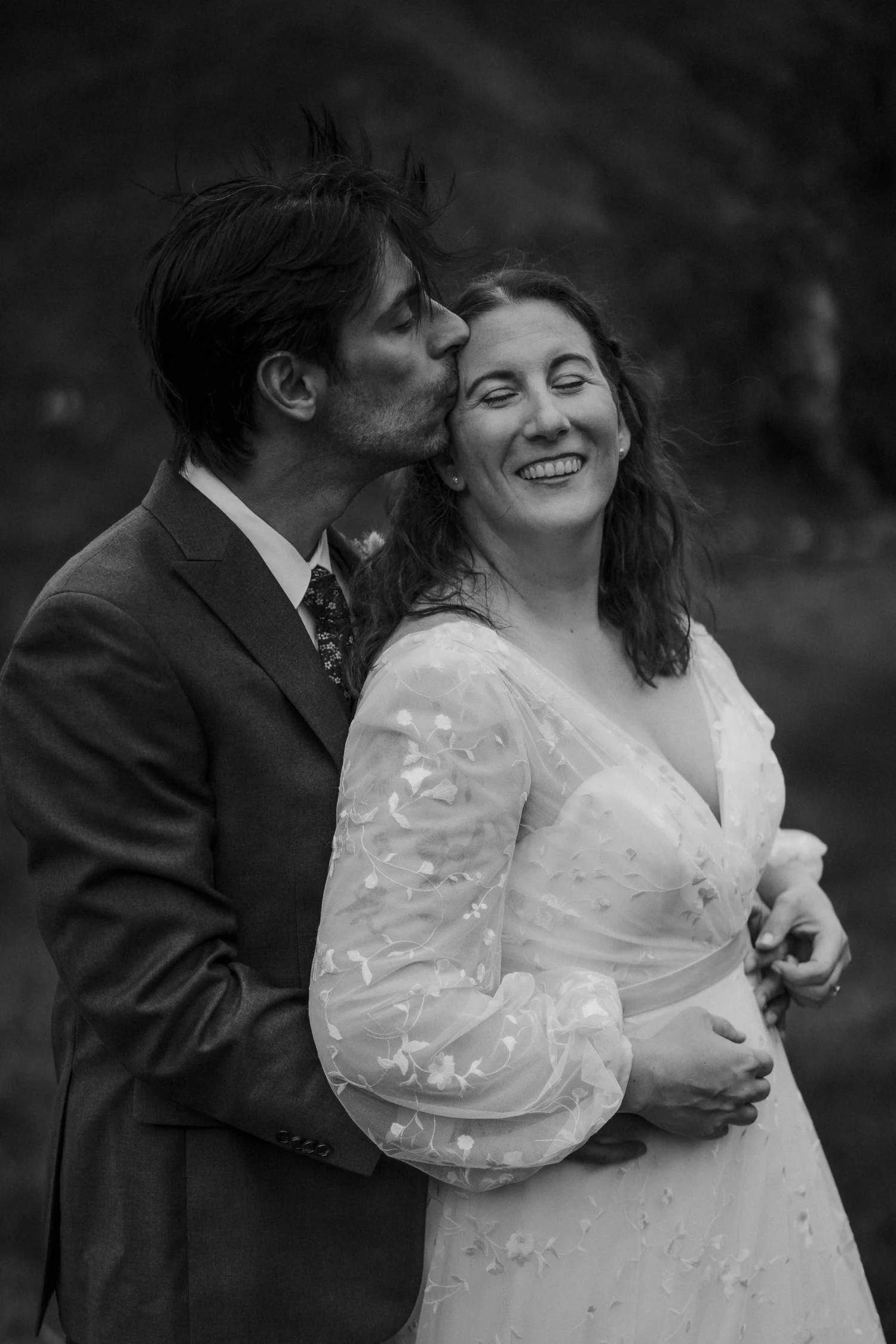 A black-and-white photo of a couple, with the man kissing the woman's temple and both smiling, in what appears to be an outdoor setting.