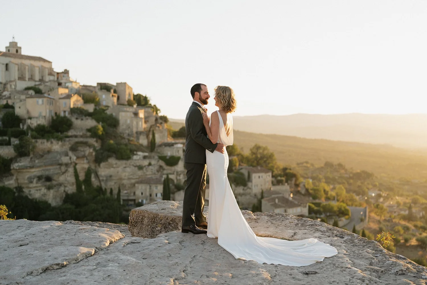 A newlywed couple stands on a rocky ledge at sunset, embracing each other with a hillside village in the background.