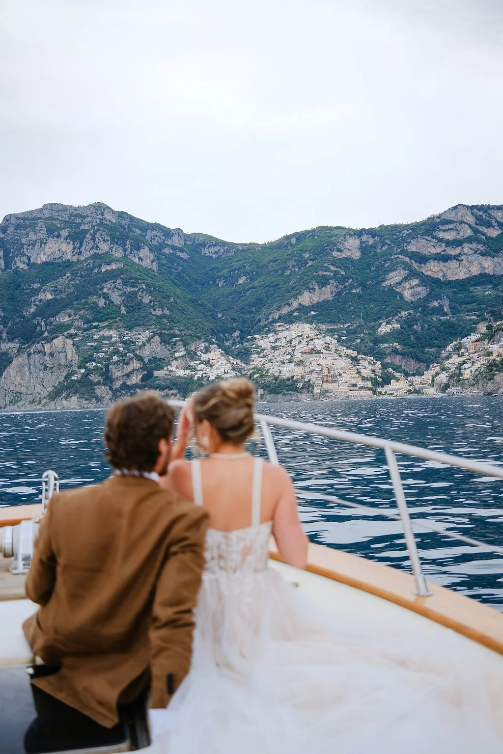 A bride and groom sitting on a boat, enjoying a scenic view of a mountain coastline with houses on the hillside.