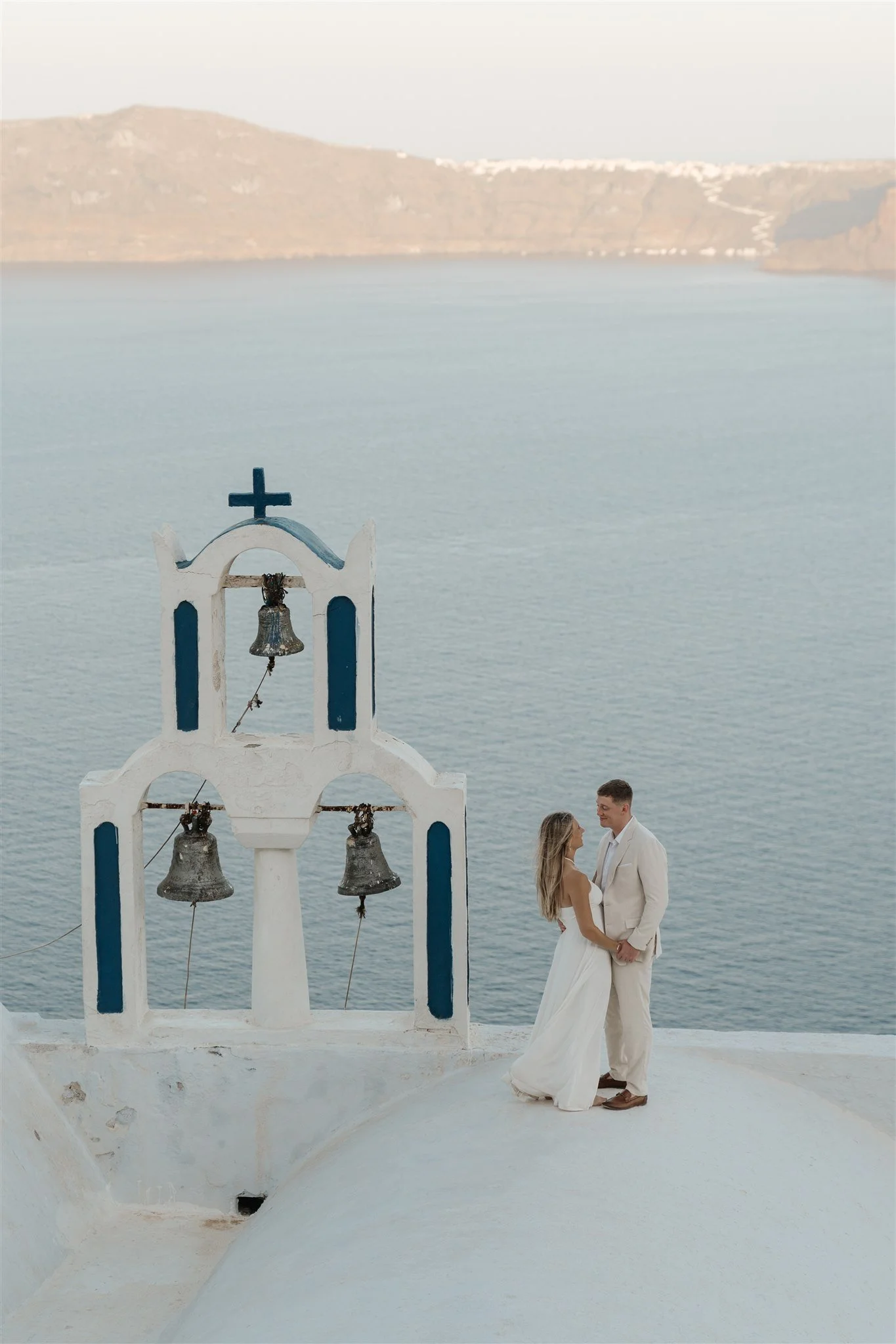 A couple dressed in wedding attire standing before a white church with bells, overlooking a calm sea with hills in the background.