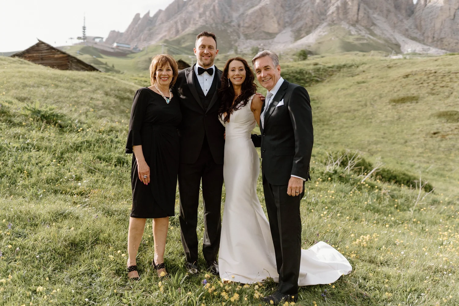 Group of four people dressed formally, standing on a grassy field with mountains and a slope with small houses in the background.