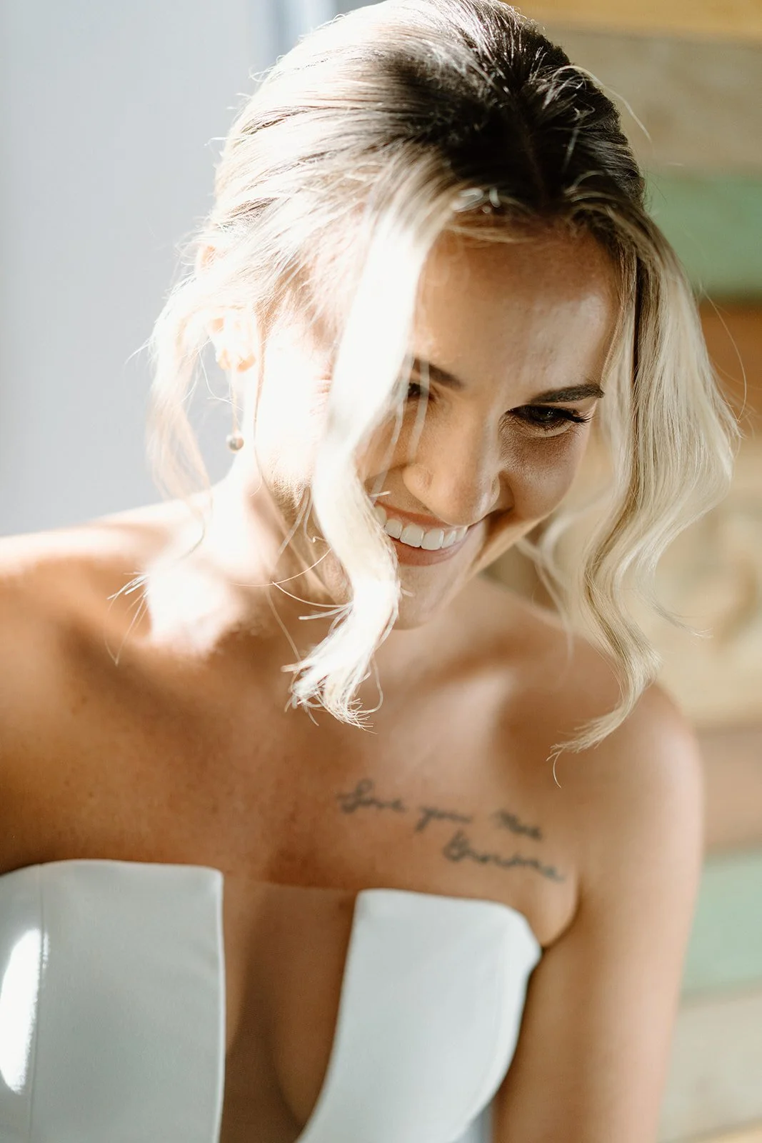 Close-up of a woman with blonde, wavy hair smiling and looking down. She has a tattoo on her chest that reads 'love you more.'