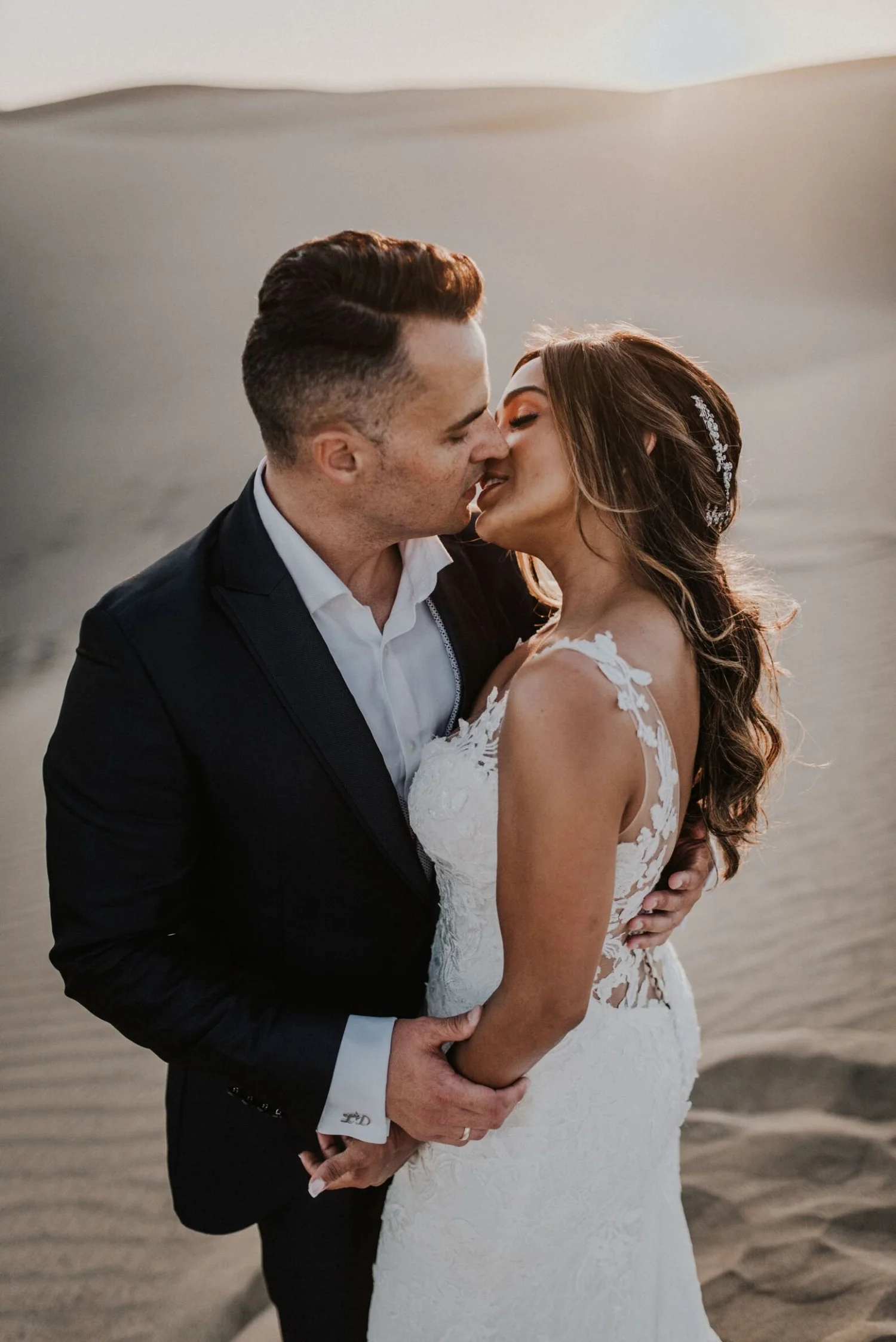 A newlywed couple sharing a kiss on the beach during sunset, with the groom in a black suit and the bride in a white lace wedding dress, holding each other closely.