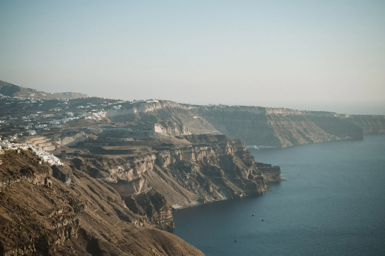 Cliffs along a coastline with houses on the upper ledges and the ocean at the bottom, hazy sky overhead.