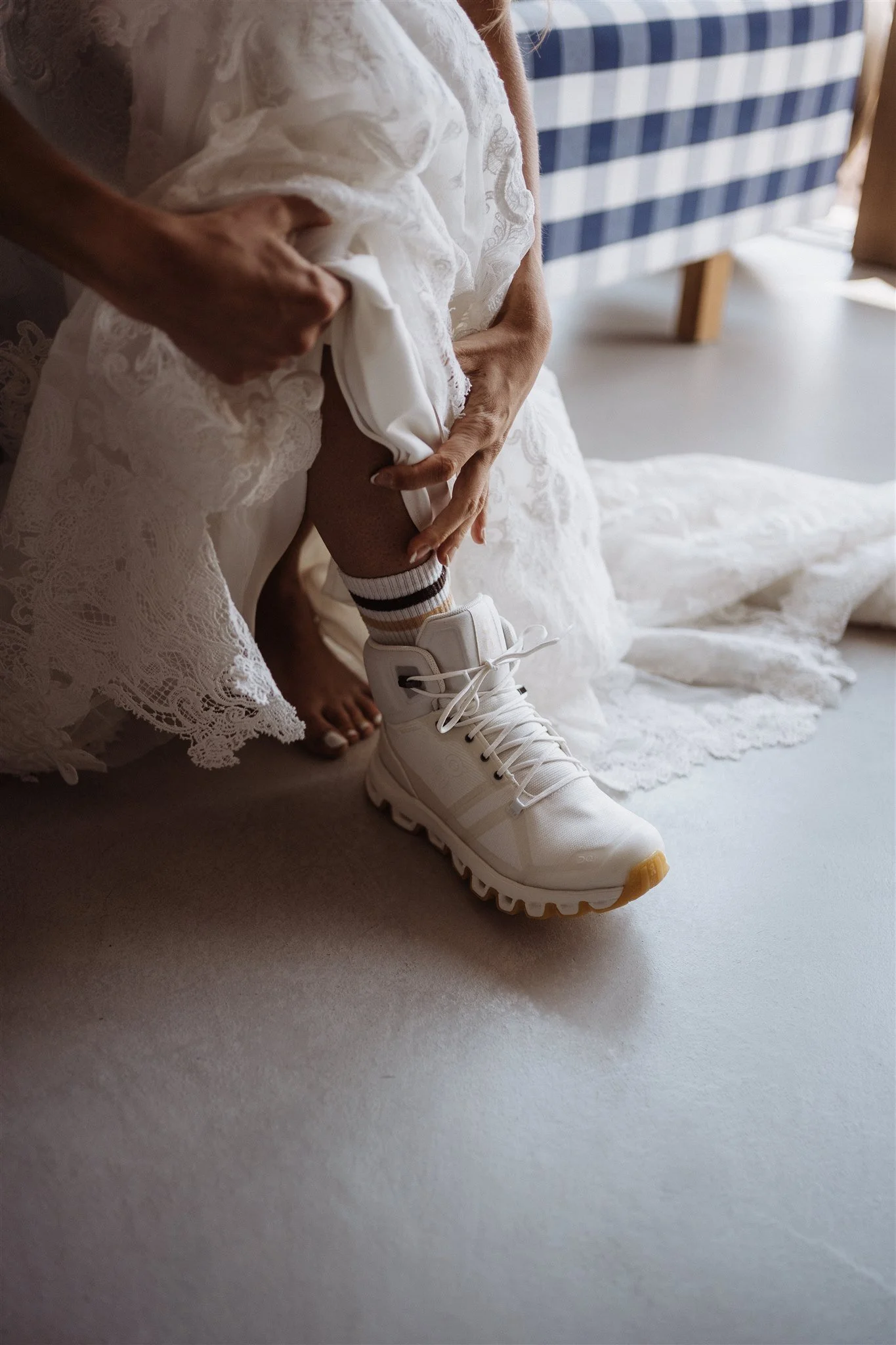 Person lacing up white athletic shoes with striped socks inside a room, with a blue and white checkered couch in the background.