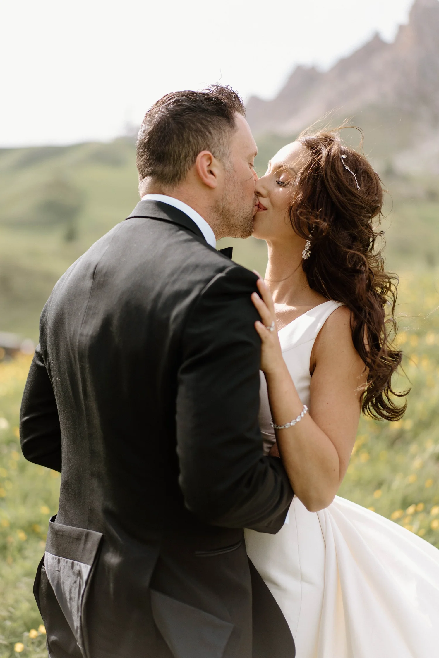 A bride and groom in a field, sharing a kiss, with mountains in the background.