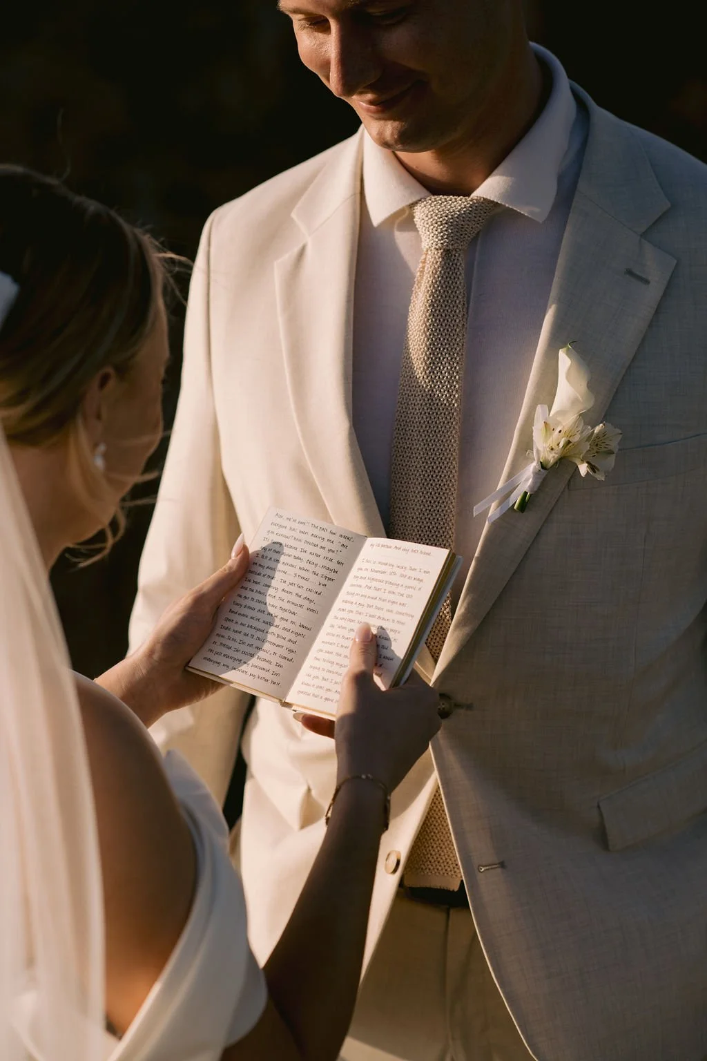 Elopement portrait of a woman holds an open book and reads to a man during a wedding ceremony. The man, dressed in a light-colored suit with a boutonnière, looks down with a smile.