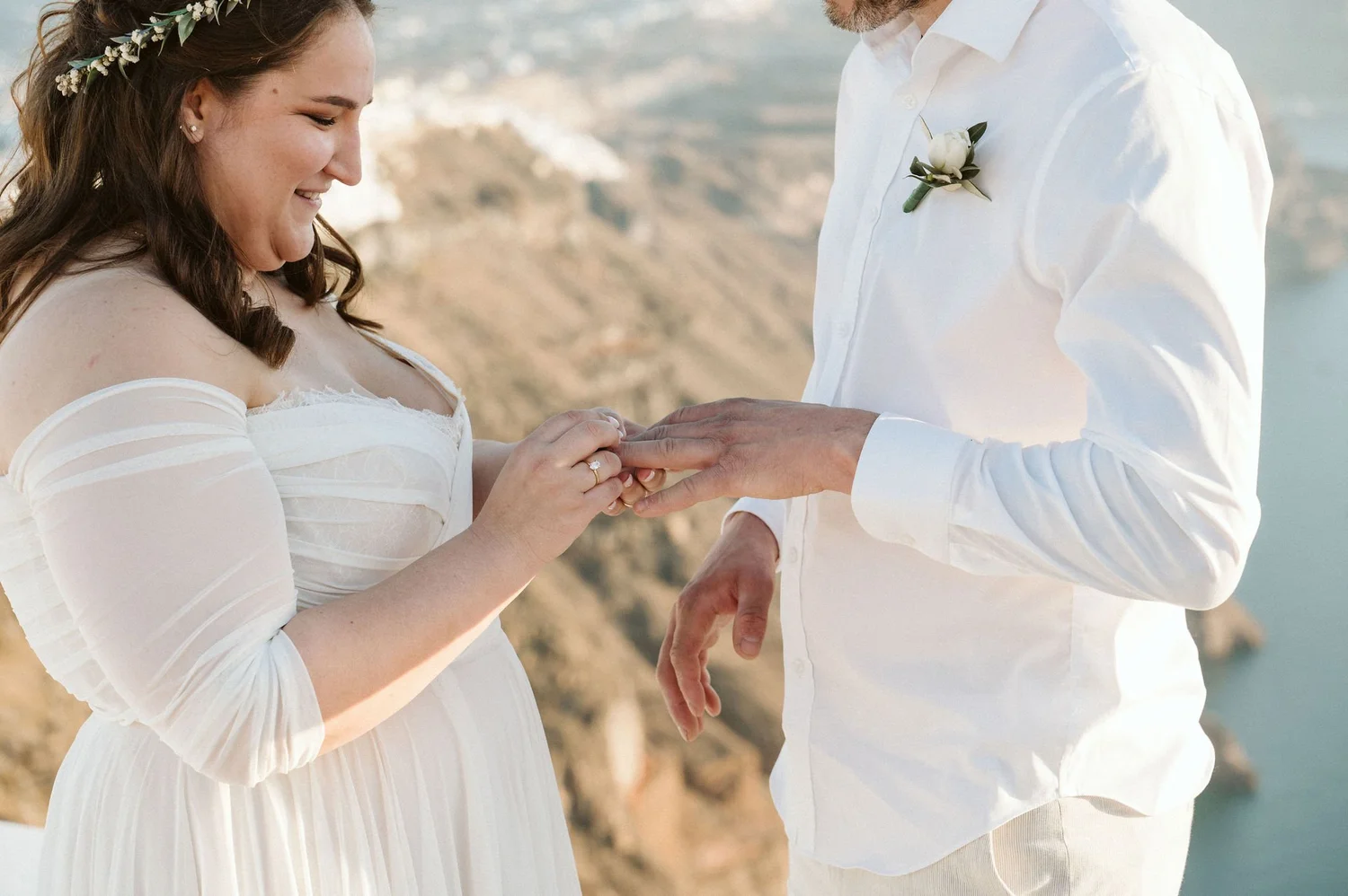 A woman with long brown hair wearing a floral headband and a white off-the-shoulder dress is holding hands with a man dressed in a white shirt and white pants. The man has a boutonniere pinned to his shirt. They are outdoors with a blurred landscape