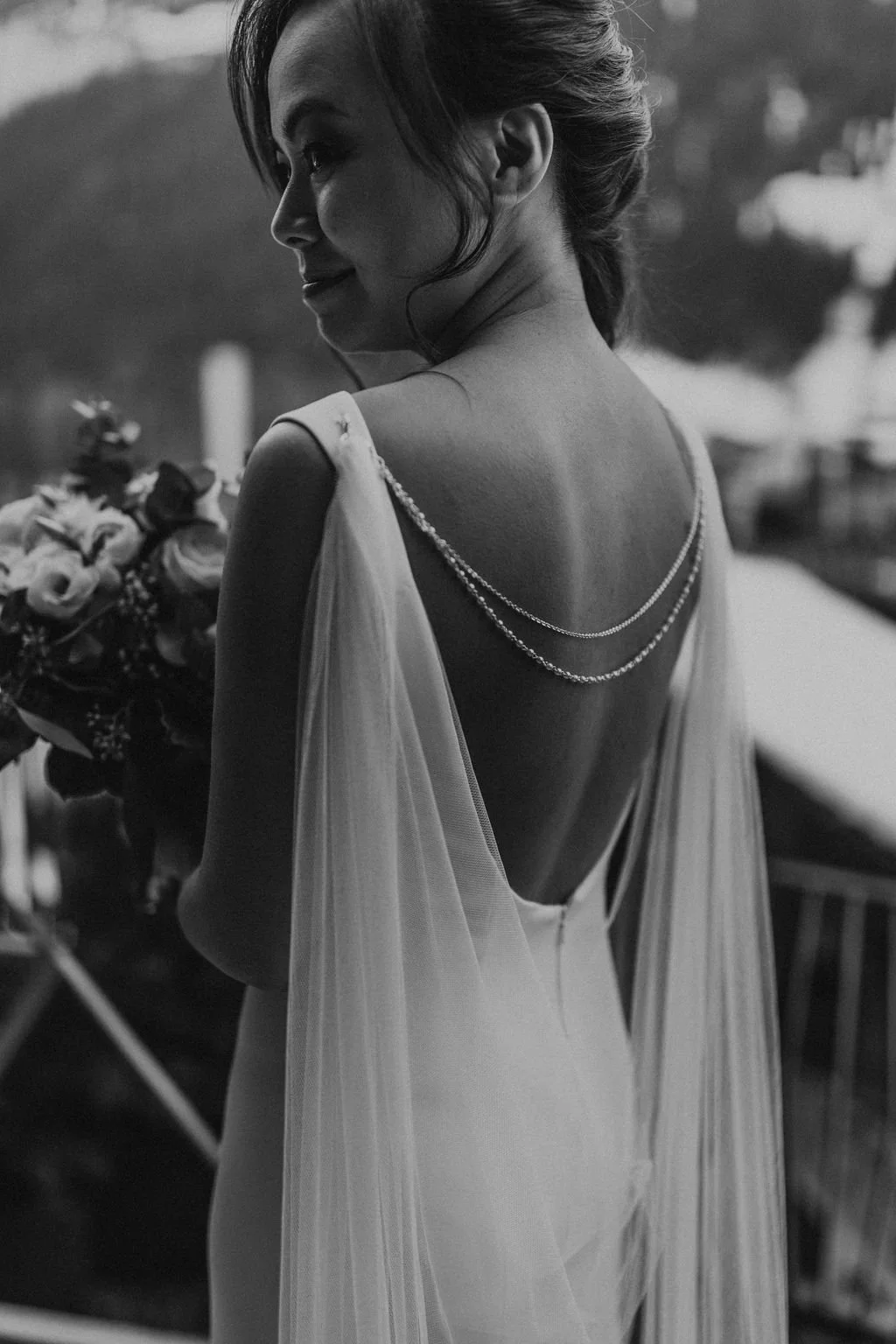 A woman in a wedding dress holds a bouquet, smiling softly and looking to the side, with focus on her back, lace details, and layered necklace, in black and white.
