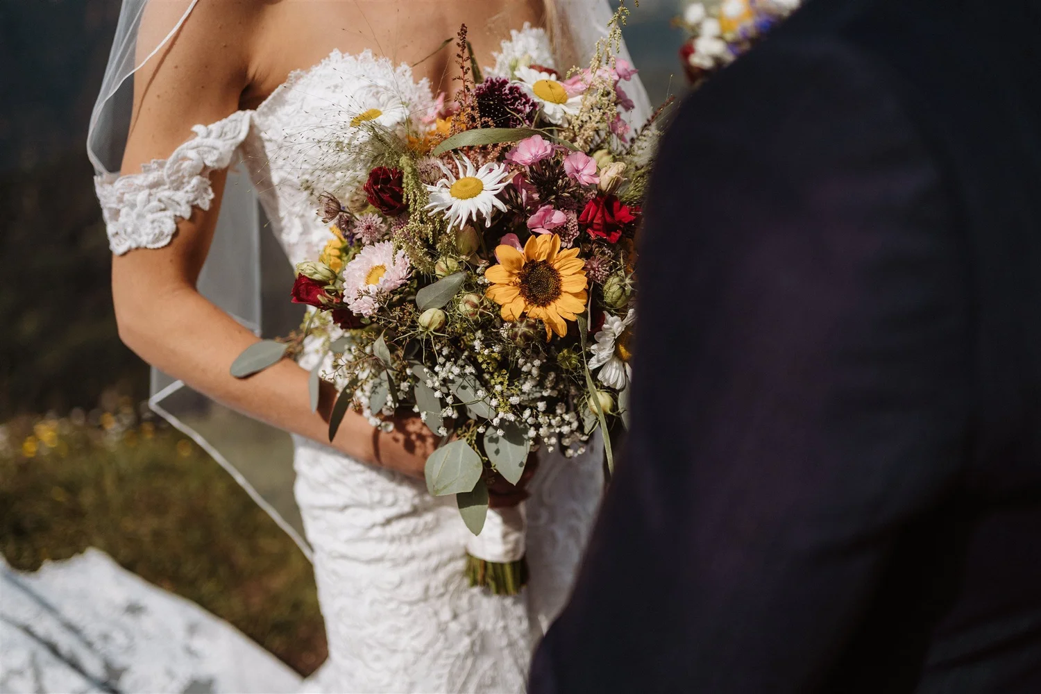 A bride holding a colorful bouquet of various flowers, including daisies, sunflowers, and other wildflowers, next to a person in a dark suit, with a blurred outdoor background.