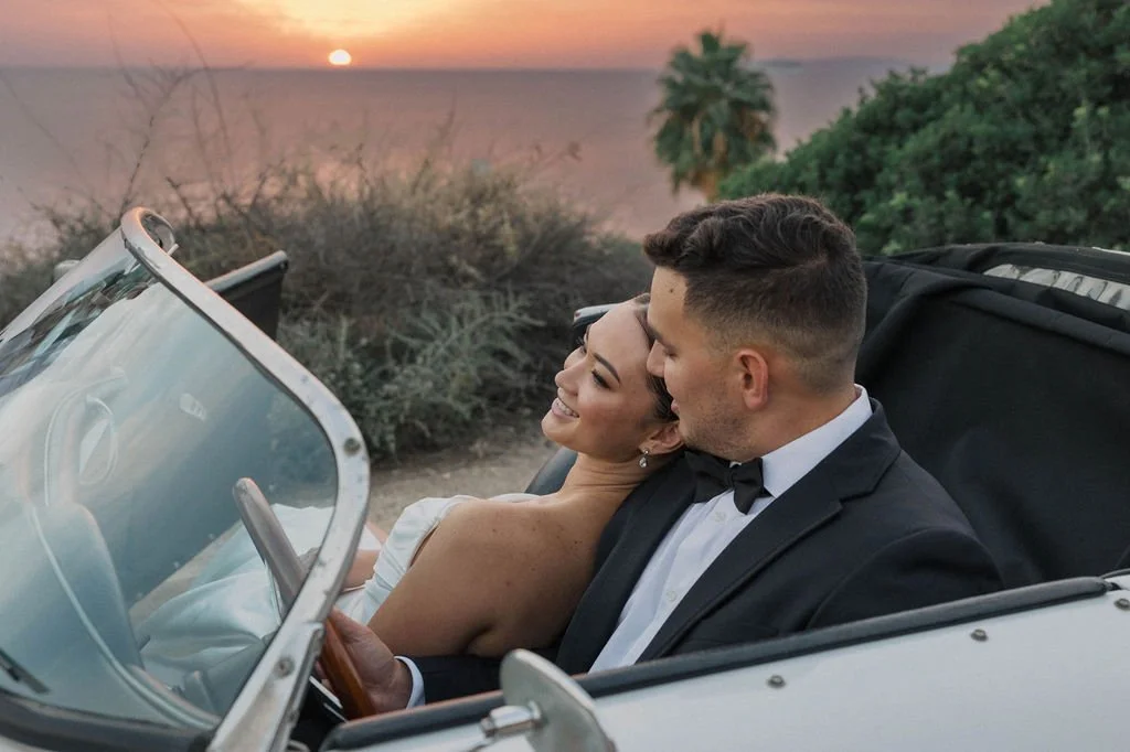 A couple in wedding attire sitting in a vintage car on a scenic overlook during sunset, with greenery and palm trees in the background.