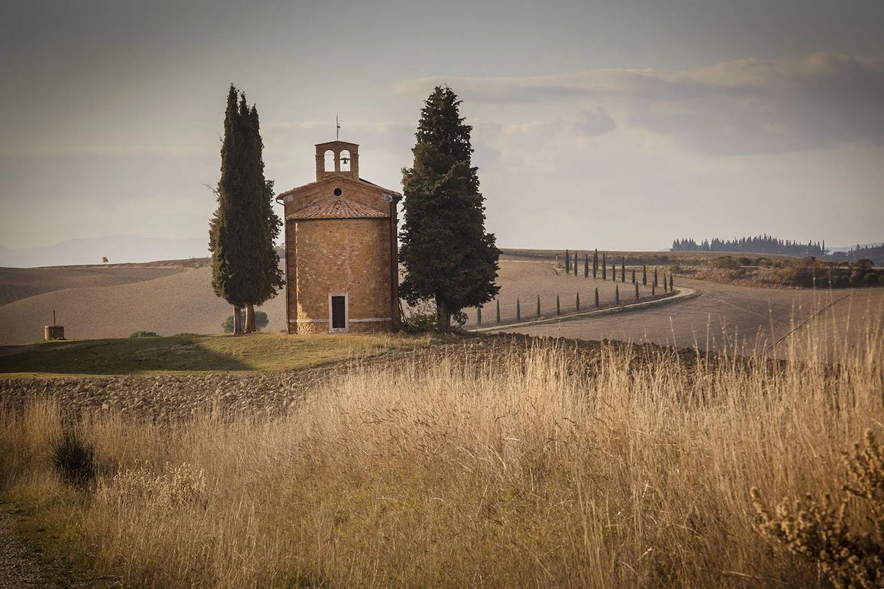 A small brick chapel with a tiled roof, surrounded by tall cypress trees, set in a rural landscape with rolling fields and a winding road in the distance.