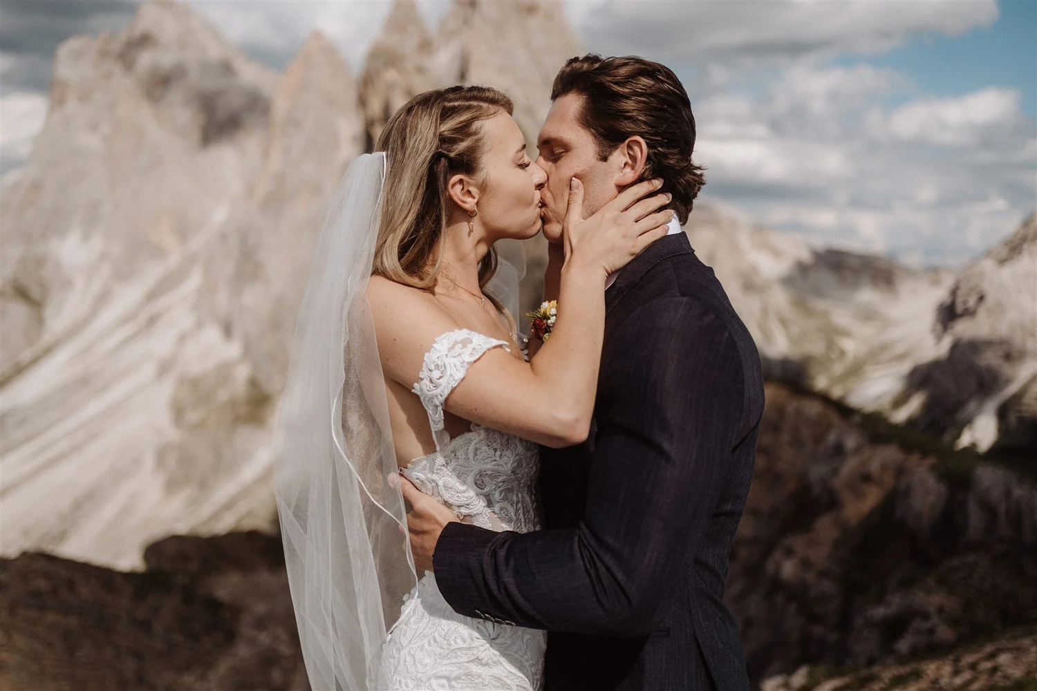 A couple kissing outdoors during their wedding, with rocky mountains and a cloudy sky in the background.