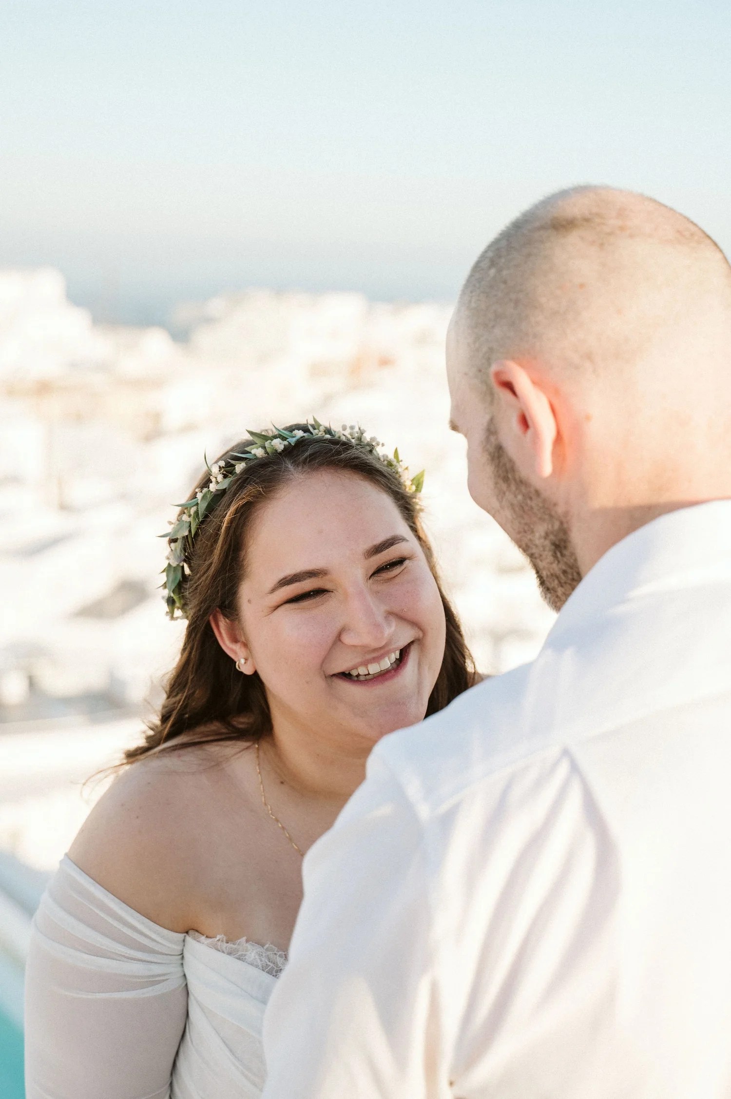 A smiling woman with long brown hair wearing a floral crown and off-the-shoulder white dress, looking at a man with a shaved head, in an outdoor setting with white buildings and blue sky in the background.