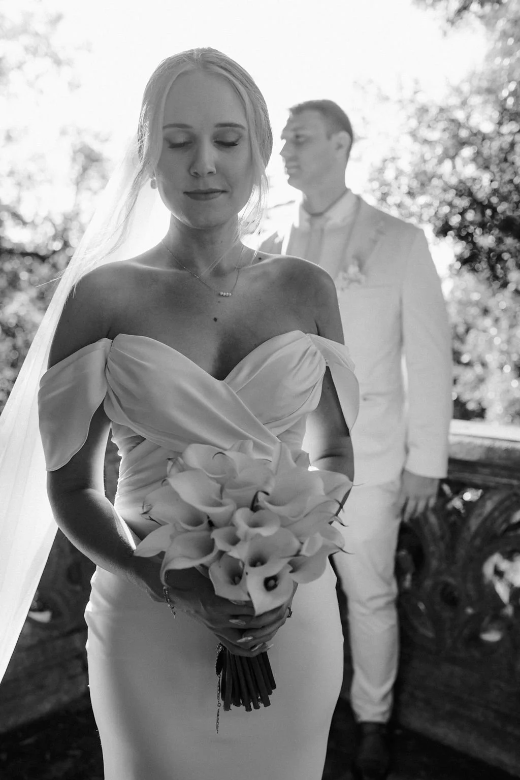 Black and white photo of a bride holding a bouquet of calla lilies with a groom in the background, outdoors with trees and sky.