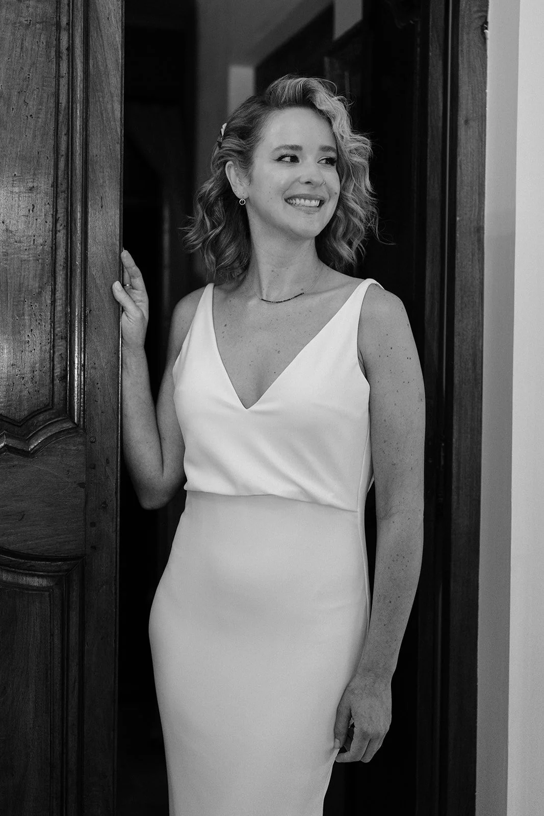A woman in a white sleeveless dress, smiling, standing in a doorway, with curly hair and earrings.